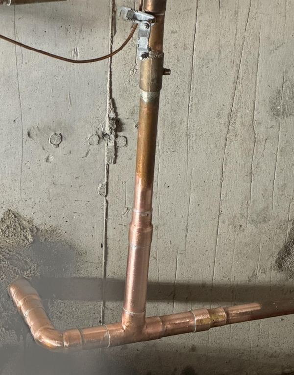 Newly soldered copper water line with a tee and an elbow, running along a concrete basement wall.
