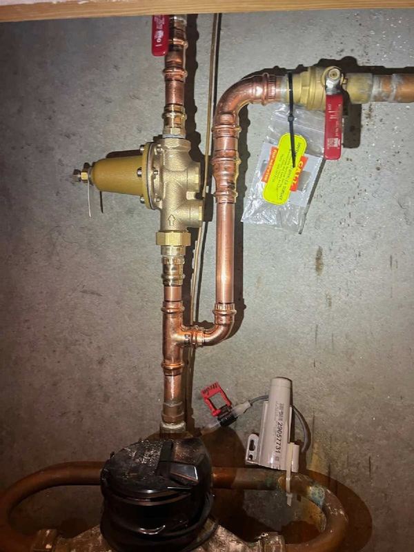 Copper water piping with a brass pressure reducing valve and red-handled ball valves against a concrete wall.