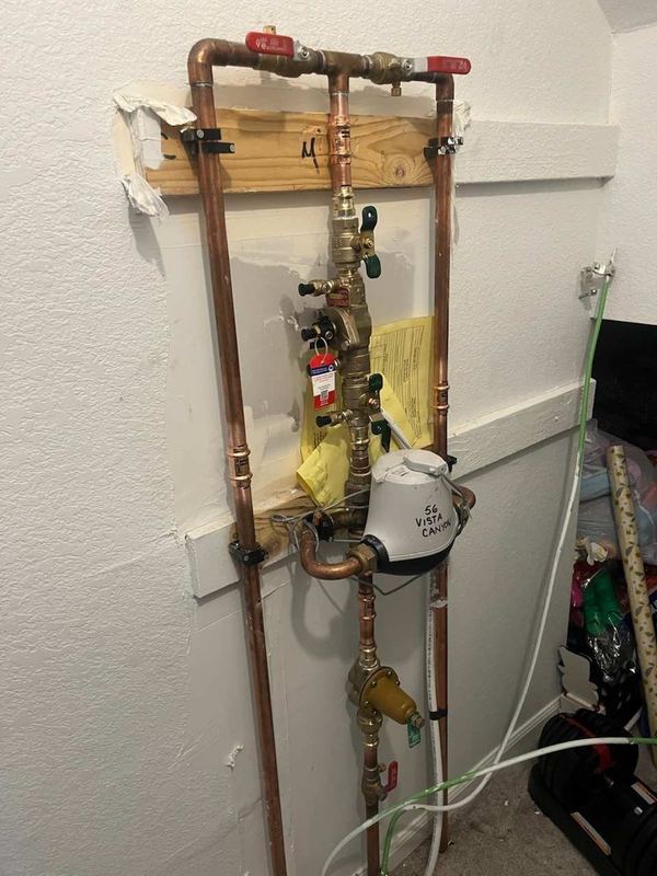 Vertical copper water piping with red-handled ball valves and brass fittings, mounted on a board against a wall.