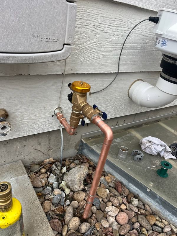 Copper line with a brass valve and fittings running up the outside wall of a house above a rock bed.