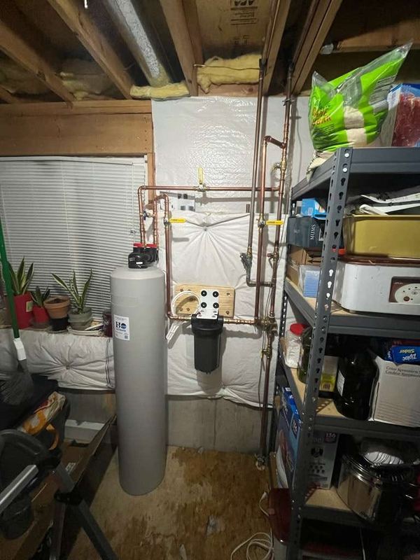 Grey whole-home water filtration tank and filter housing connected to copper piping in an unfinished basement.