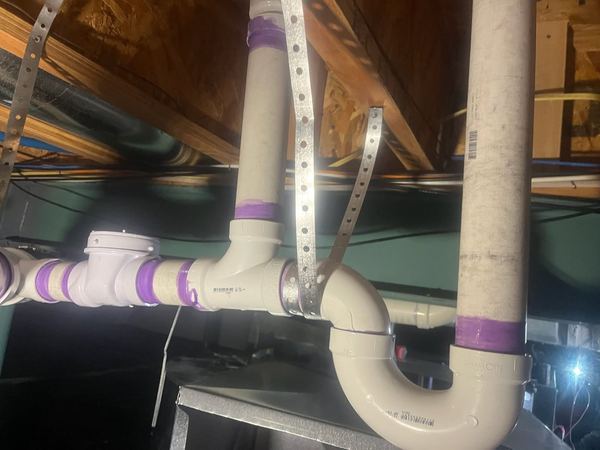 White PVC drain and vent piping with purple primer marks, hung from metal straps along a basement ceiling.
