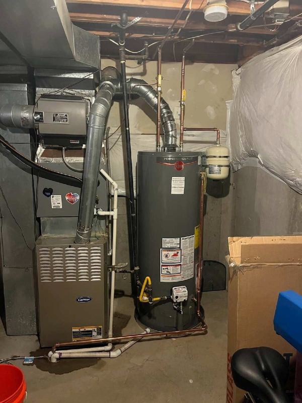 Tank water heater installed next to a furnace in an unfinished basement, with copper and PVC piping.