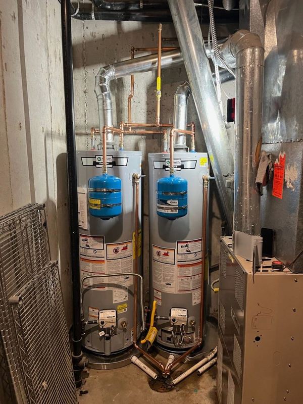 Two tank water heaters side by side in a utility closet, each with a blue expansion tank and copper piping overhead.