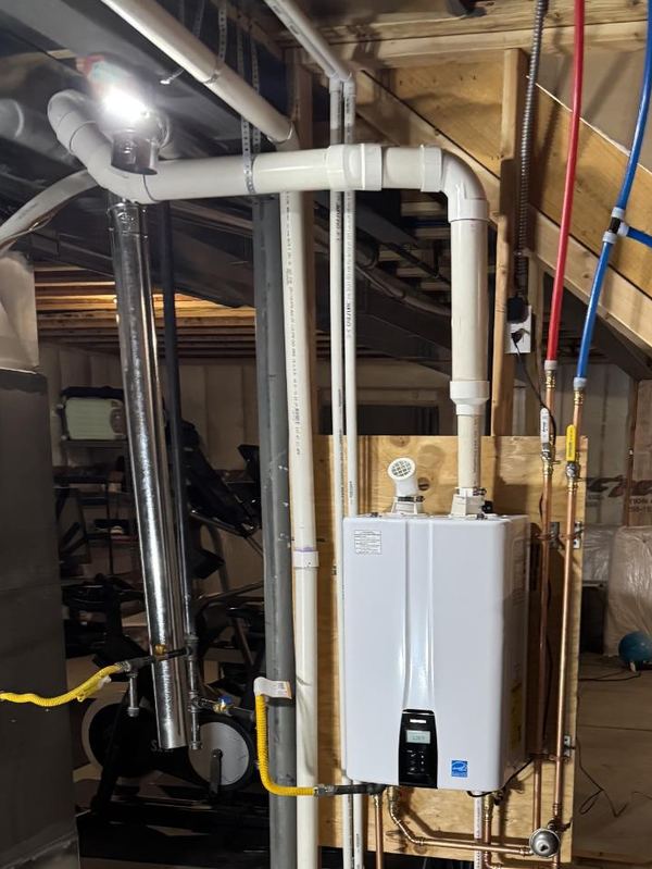White wall-mounted tankless gas water heater in an unfinished basement, with copper water connections and a yellow gas line below, white PVC vent pipes and red and blue PEX lines overhead.