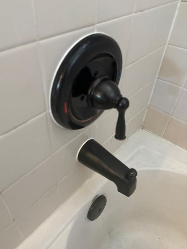 Oil-rubbed bronze tub and shower valve trim with a single lever handle above a matching tub spout on a white tiled wall.