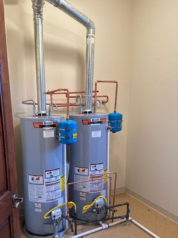 Two gas tank water heaters installed side by side in a utility room, each with a blue thermal expansion tank, copper water piping and metal vent pipes rising to the ceiling.
